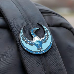 May include: A blue and black enamel pin featuring a stylized loon design with geometric patterns and a crescent moon shape. The pin is attached to a black backpack strap. The loon has a blue body, white chest, and a dark blue beak.