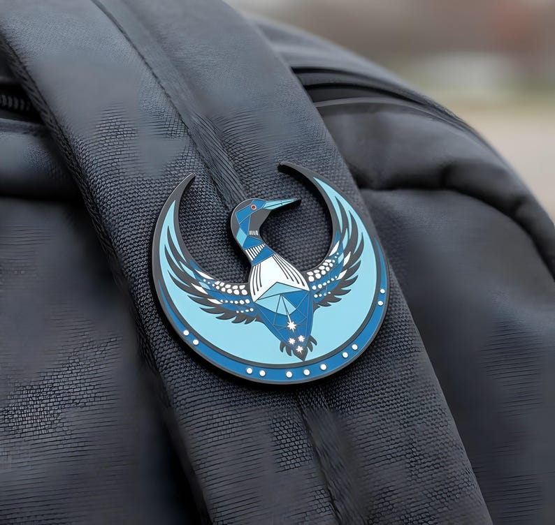 May include: A blue and black enamel pin featuring a stylized loon design with geometric patterns and a crescent moon shape. The pin is attached to a black backpack strap. The loon has a blue body, white chest, and a dark blue beak.