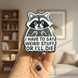 May include: A black and white glittery acrylic badge reel featuring a cartoon raccoon illustration. The text reads: I HAVE TO SAY WEIRD STUFF OR I'LL DIE. The design is attached to a clear plastic strap with a circular button fastener at the bottom.
