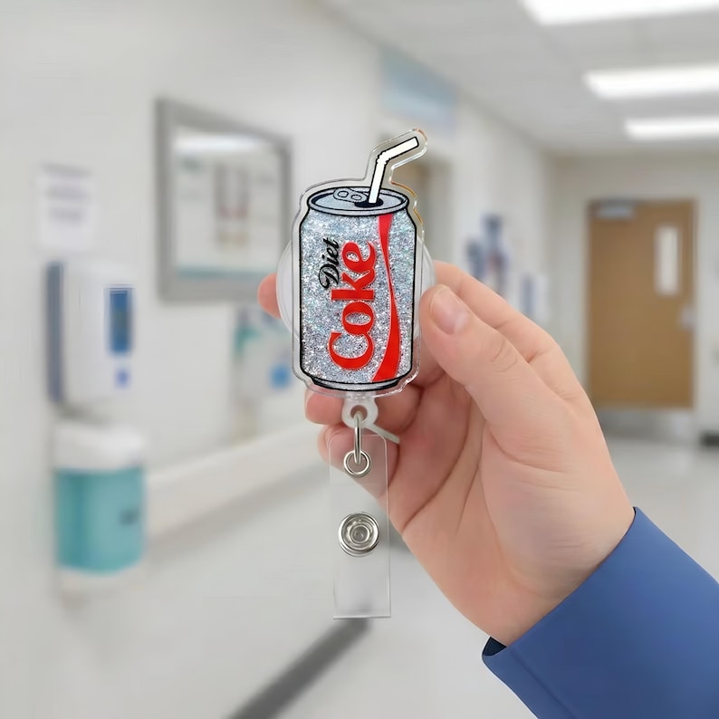 May include: A Diet Coke badge reel with a silver glitter background and red lettering. The badge reel is in the shape of a Diet Coke can with a white straw. The reel has a clear plastic strap and a metal clip.