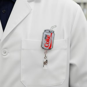 May include: A Diet Coke badge reel featuring a silver glitter can design with a red outline. The reel is attached to a white pocket on a white lab coat.
