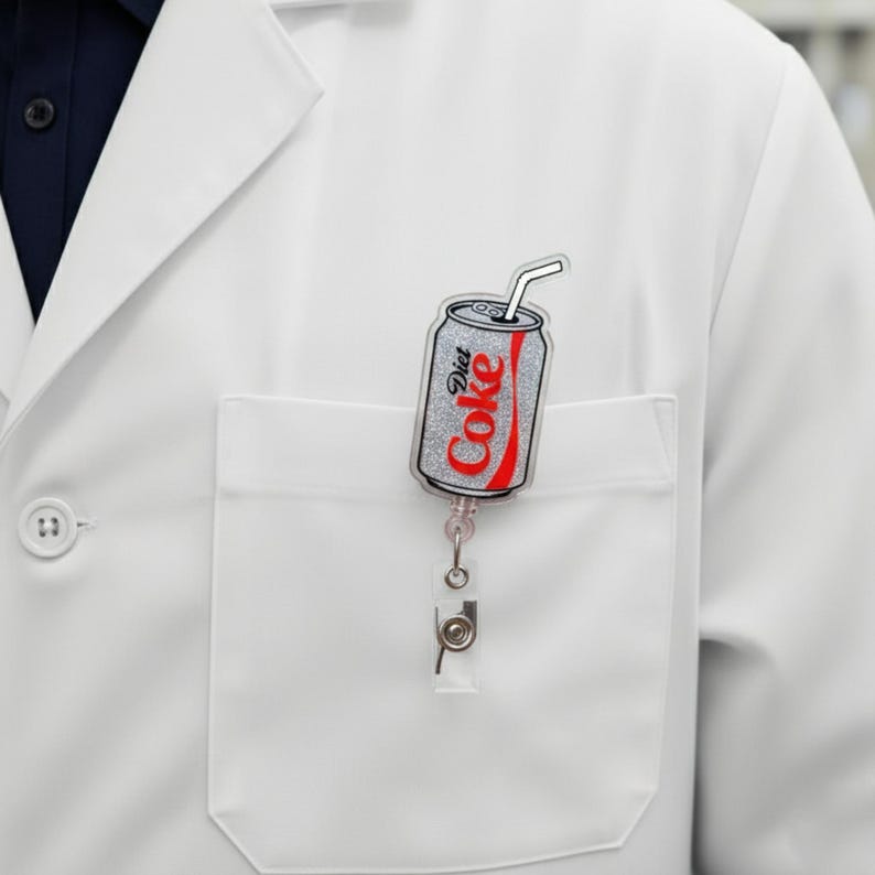 May include: A Diet Coke badge reel featuring a silver glitter can design with a red outline. The reel is attached to a white pocket on a white lab coat.