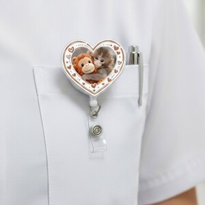 May include: A white heart-shaped badge reel with a brown border and a cartoon image of two monkeys hugging. The badge reel is attached to a white shirt pocket with a pen.