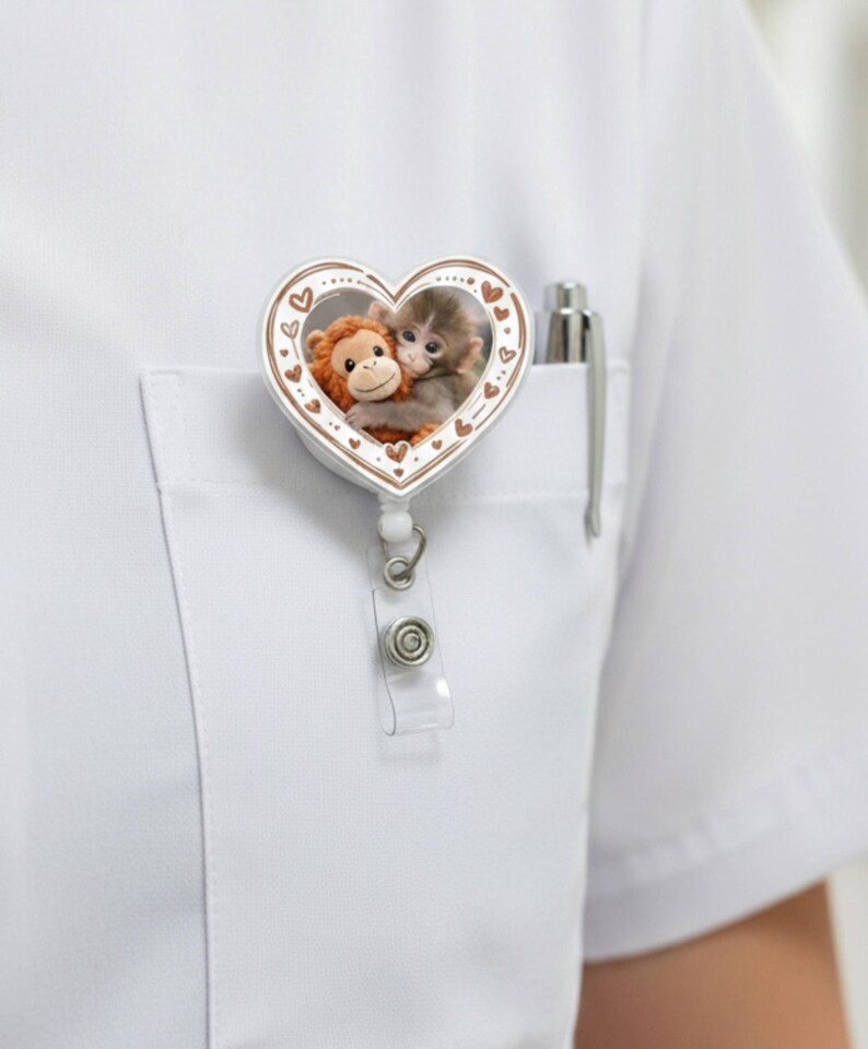 May include: A white heart-shaped badge reel with a brown border and a cartoon image of two monkeys hugging. The badge reel is attached to a white shirt pocket with a pen.