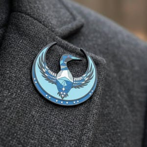 May include: A round, blue and black pin featuring a stylized bird design. The bird has outstretched wings and is set against a crescent moon shape. The pin is attached to a gray fabric.