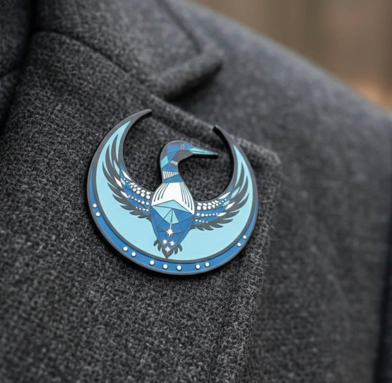 May include: A round, blue and black pin featuring a stylized bird design. The bird has outstretched wings and is set against a crescent moon shape. The pin is attached to a gray fabric.