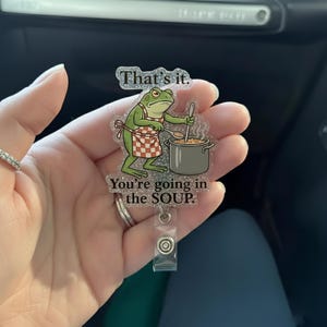 May include: A clear, glittery badge reel featuring a cartoon frog wearing a red and white checkered apron, stirring a pot of soup. Text above reads "That's it." and below "You're going in the SOUP."