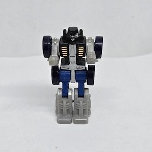 以下が含まれることがあります： 立っている小さなロボットのおもちゃのフィギュア。ロボットは黒い頭、金色のアクセントが付いた灰色の胴体、青い脚をしています。4つの黒い車輪と灰色の腕と足があります。おもちゃは白い背景に設定されています。