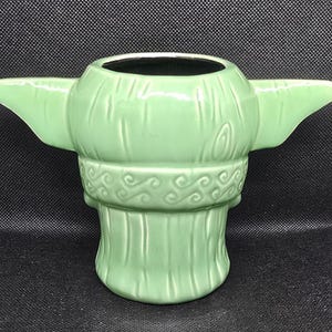 May include: A light green ceramic mug shaped like a character's head with large ears extending from the sides. The mug has a wide opening and a detailed design, including textured lines and decorative swirls around the neck.