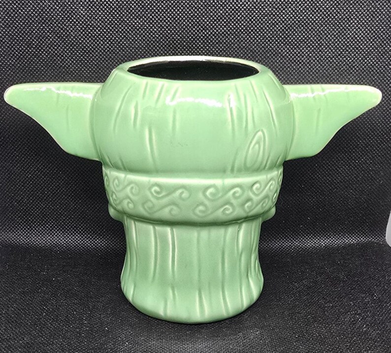 May include: A light green ceramic mug shaped like a character's head with large ears extending from the sides. The mug has a wide opening and a detailed design, including textured lines and decorative swirls around the neck.