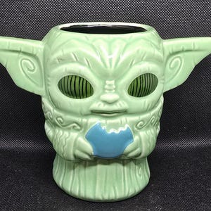 May include: A light green ceramic mug shaped like a character with large ears and large eyes. The mug has a blue accent on the front and is designed with a textured surface. The mug is a collectible item.