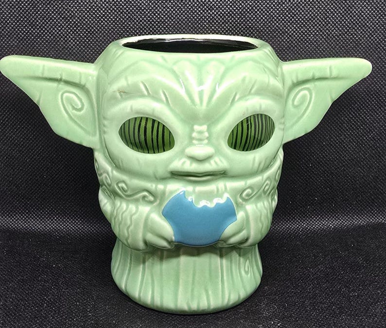 May include: A light green ceramic mug shaped like a character with large ears and large eyes. The mug has a blue accent on the front and is designed with a textured surface. The mug is a collectible item.