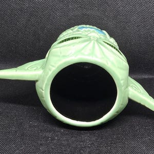 May include: A light green ceramic mug shaped like a character's head, with large ears extending from the sides. The mug has a large, open mouth and a black interior. The character's head has a textured surface and blue accents.