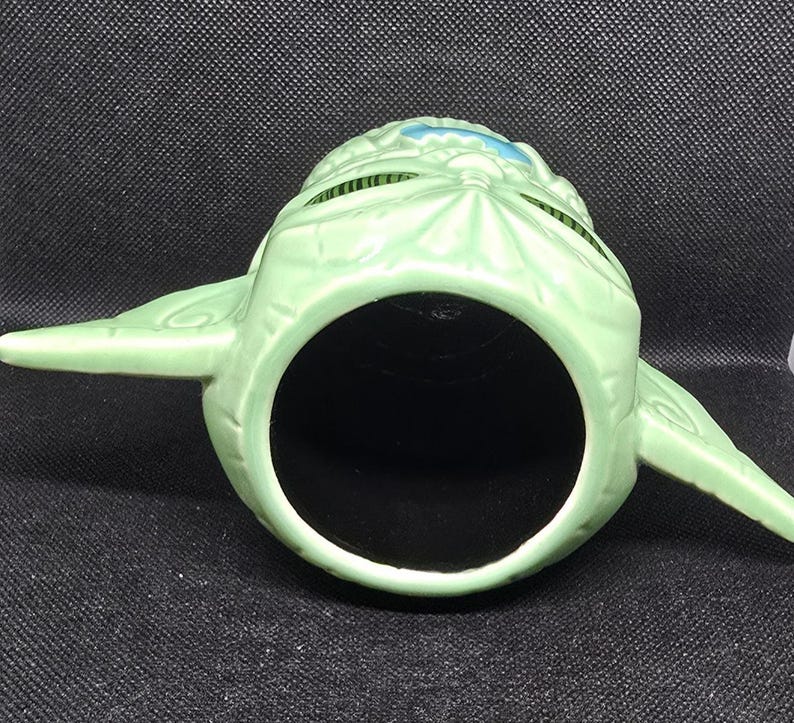 May include: A light green ceramic mug shaped like a character's head, with large ears extending from the sides. The mug has a large, open mouth and a black interior. The character's head has a textured surface and blue accents.
