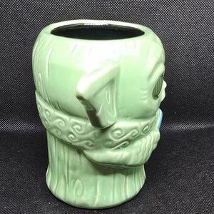 May include: A light green ceramic mug shaped like a character's head, featuring large ears and detailed facial features. The mug has a wide opening and a handle-like ear. The design includes textured details and a light blue accent.