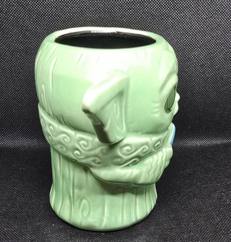 May include: A light green ceramic mug shaped like a character's head, featuring large ears and detailed facial features. The mug has a wide opening and a handle-like ear. The design includes textured details and a light blue accent.