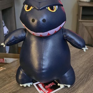 May include: A navy blue Godzilla plush toy with yellow and black eyes, a red and white mouth, and white claws. The plush toy is sitting on a wooden surface. A tag with the word "Godzilla" is visible.