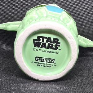 May include: A light green ceramic mug shaped like a character from Star Wars, with large ears and a blue accent. The bottom of the mug features the Star Wars logo, Geek Tiki branding, and copyright information. Made in China.