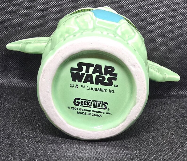 May include: A light green ceramic mug shaped like a character from Star Wars, with large ears and a blue accent. The bottom of the mug features the Star Wars logo, Geek Tiki branding, and copyright information. Made in China.