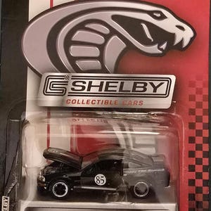 May include: A black 2007 Shelby GT collectible car model with the hood open, displayed in a clear plastic case. The packaging features the Shelby logo and a warning about small parts. The car has the number 85 on the side.