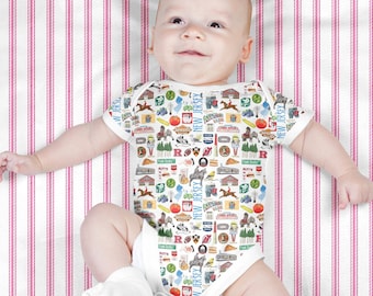 May include: A white baby onesie with a colorful pattern of New Jersey icons, including the state flag, a horse, a tomato, and a slice of pizza. The baby is lying on a pink and white striped blanket.