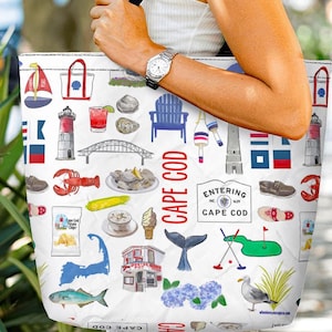 May include: White tote bag featuring a Cape Cod theme with illustrations of a lighthouse, lobster, bridge, and other coastal elements. The bag has black straps and is held by a person.