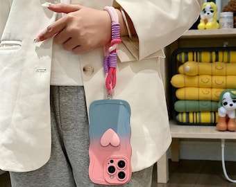 Op de afbeelding: Een roze en blauwe ombre telefoonhoesje met een hartontwerp, bevestigd aan een veelkleurig koord. De hoes heeft een golvende rand en een camera-uitsparing. Het koord is roze, paars en rood.
