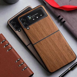 Puede incluir: Dos fundas de teléfono con textura de madera en tonos marrones oscuros. Una funda muestra una interfaz de reproductor de música. Las fundas tienen un borde negro y la palabra "AIORIA" está impresa en la esquina inferior derecha. Sobre una superficie gris claro.