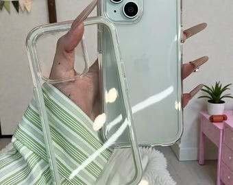 May include: Clear phone case with a transparent design. The case is shown next to a light green phone. The phone has a dual-lens camera and a silver Apple logo. The case is held in front of a green and white striped shirt.