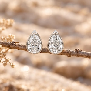 May include: A pair of pear-shaped stud earrings featuring clear, faceted stones set in a metallic frame with a beaded edge. The earrings are positioned side-by-side, resting on a thin, textured brown twig against a soft, out-of-focus background.