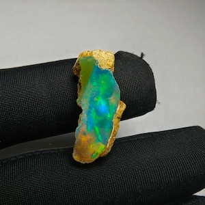 Puede incluir: Una piedra preciosa de ópalo en bruto con colores vibrantes de verde, azul y amarillo. El ópalo presenta una textura natural y rugosa, con un juego de colores iridiscentes. La piedra es un espécimen natural único, sobre un fondo neutro.