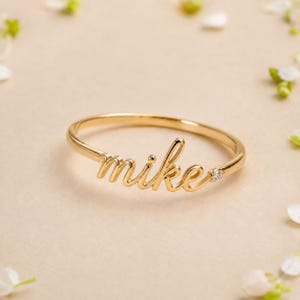 Puede incluir: Un anillo de tono dorado que presenta el nombre mike en una fuente cursiva. La banda es delgada y pulida, con una pequeña piedra preciosa transparente de corte redondo engastada en el metal justo a la derecha del nombre, añadiendo brillo.