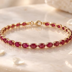 Puede incluir: Una pulsera de cadena dorada con una hilera continua de rubíes de color rojo intenso en talla ovalada, engastados individualmente en garras de oro. La pulsera se asegura con un cierre de mosquetón dorado, creando un diseño circular.