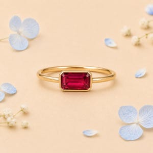May include: A gold ring featuring a horizontal, emerald-cut, deep pink gemstone set in a bezel setting. The band is thin and polished, showcasing a clean, minimalist design that highlights the vibrant colour and rectangular shape of the centre stone.