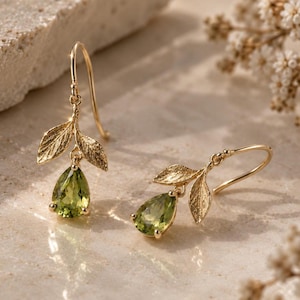 May include: A pair of gold drop earrings featuring two textured leaf-shaped charms and a single pear-cut green gemstone suspended from a curved hook. The metal has a matte finish, and the green stones are faceted to reflect light clearly.