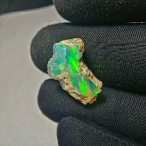May include: A rough-cut opal gemstone with vibrant green and yellow internal colors. The opal is set against a dark background, highlighting its iridescent qualities. The stone has a natural, unpolished appearance.