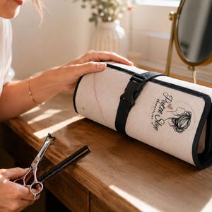 Op de afbeelding: Een beige oprolbare gereedschapsorganizer met zwarte randen en een zwarte plastic gespriem. De stof is voorzien van een zwart grafisch logo van een persoon met een opgestoken kapsel en de tekst Peyton Skye Hairstylist. Haarschaar en kam aanwezig.
