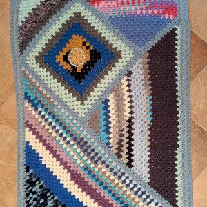 May include: A handmade, colorful crochet blanket with a geometric design. The blanket features a variety of colors, including blues, greens, purples, and browns. The blanket has a light blue border and measures approximately 40 x 50 inches.