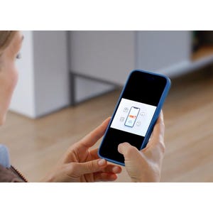 May include: A person holding a blue smartphone displaying an app interface with a phone icon and other symbols. The phone has a black screen and a blue case. The background is blurred, showing a room.