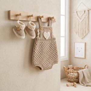 May include: A light brown crochet romper featuring a white heart applique and two wooden buttons on the straps, hanging on a wooden wall rack alongside a pair of matching light brown and cream crochet booties with small wooden button accents.