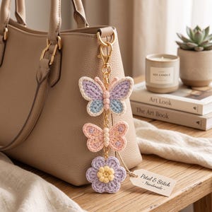 May include: A crochet bag charm featuring two butterflies and one flower in pastel shades of lavender, light blue, and soft pink, attached to a gold-tone metal clasp. A small white paper tag attached to the charm reads Petal & Stitch - Handmade.