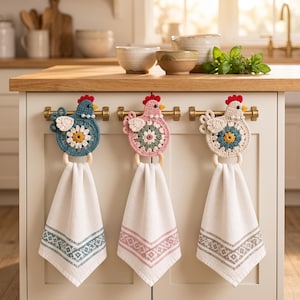 Puede incluir: Tres soportes para toallas de ganchillo con forma de gallina colgados de un tirador de armario. Cada uno presenta un cuerpo de granny square colorido, una cresta roja, un pico amarillo y un anillo de madera que sujeta una toalla blanca.