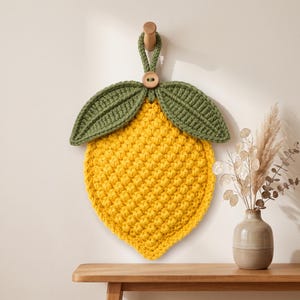 Puede incluir: Un tapiz de crochet con forma de limón que presenta un cuerpo texturizado de color amarillo brillante con un patrón de punto de burbuja. Dos hojas de crochet verde oscuro están unidas en la parte superior, centradas por un pequeño botón de madera.