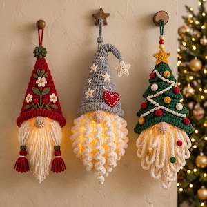 May include: Three hanging crochet gnome ornaments with internal lights. The left gnome has a red hat with white flowers, the centre gnome has a grey hat with stars and a red heart, and the right gnome has a green tree-shaped hat with colourful pom-poms.