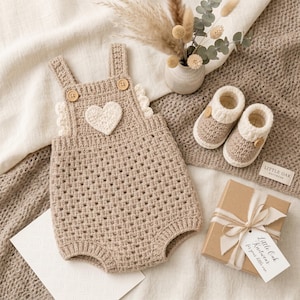 May include: A beige crochet romper featuring a white heart appliqu&eacute; on the chest, two wooden buttons on the straps, and ruffled side details. Included are matching beige and white crochet booties and a gift box with a tag reading Little Oak Knitwear.