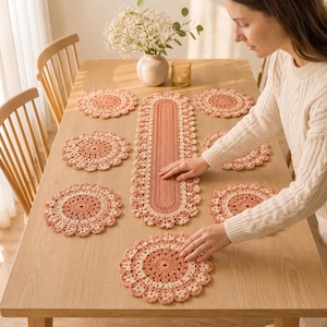 Puede incluir: Un juego de mantelería de ganchillo que incluye un camino de mesa ovalado largo y seis manteles individuales circulares a juego. Cada pieza presenta un diseño bicolor en tonos terracota y crema, con intrincados patrones florales.