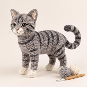 May include: A gray and white crocheted cat figurine with dark gray tabby stripes, brown eyes, and a pink nose. The cat stands upright with a curved tail. Beside the cat are two small balls of yarn in gray and cream, and a wooden crochet hook.
