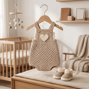 May include: A beige crochet baby romper featuring a white heart appliqu&eacute; on the chest, two wooden buttons on the straps, and a textured open-stitch pattern. The romper is paired with matching beige and white crochet booties with wooden buttons.
