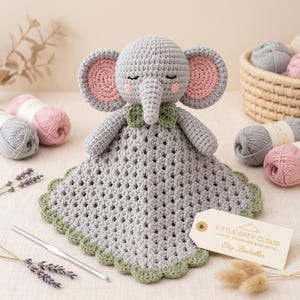 May include: A grey crochet elephant lovey with pink inner ears, a green bow tie, and a scalloped green border. A white tag attached to the toy reads: LITTLE GREY CLOUD - LUXURY HANDMADE BABY GIFTS - Etsy Bestseller. The toy is made of yarn.