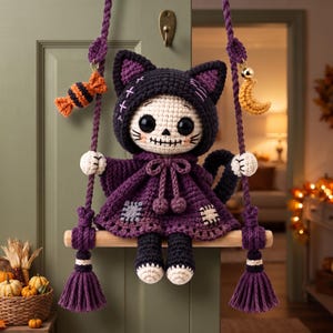 Könnte beinhalten: Eine gehäkelte Puppe mit totenkopfartigem Gesicht und schwarzen Katzenohren, die einen dunkelvioletten Kapuzenumhang mit Flicken trägt. Die Puppe sitzt auf einer Holzschaukel, die an violetten Seilen hängt und mit einem orangefarbenen Bonbon verziert ist.